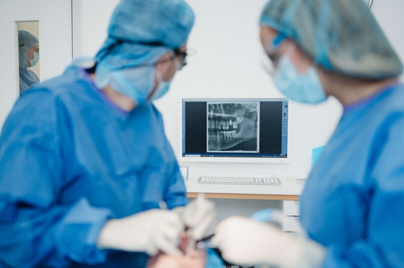 Dr. Will und Dr. Mai im OP-Saal in OP-Kleidung. Im Hintergrund ist ein Bildschirm mit einem R&ouml;ntgenbild eines Kiefers zu sehen.
