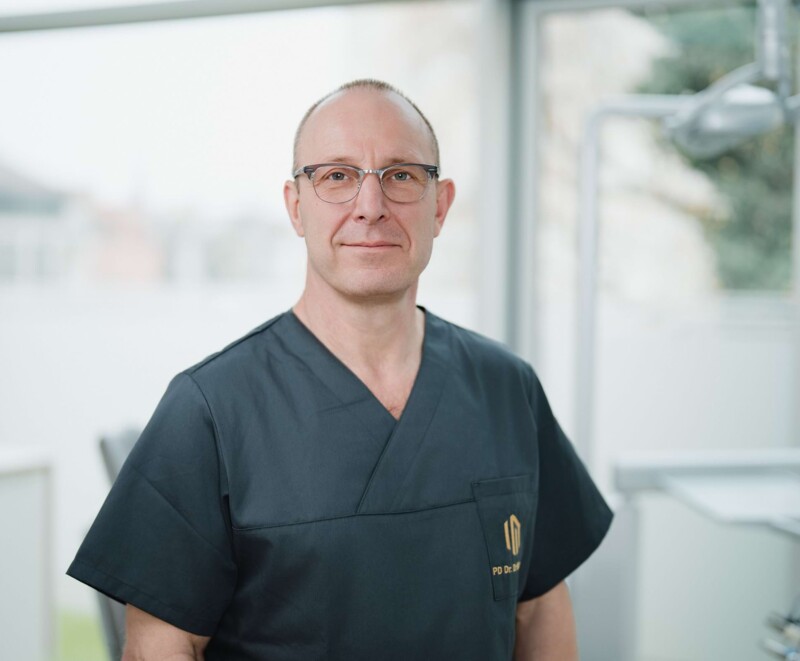 Freundliches Portrait von PD Dr. Dr. Klaus-Kristian W&uuml;rzler in schwarz/grauem Kasack vor der Fensterfront eines Behandlungszimmers