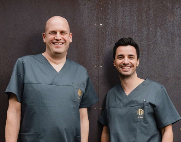 Dr. Florian Will und Dr. Alexander Mai lachen vor dem Praxisgeb&auml;ude in die Kamera