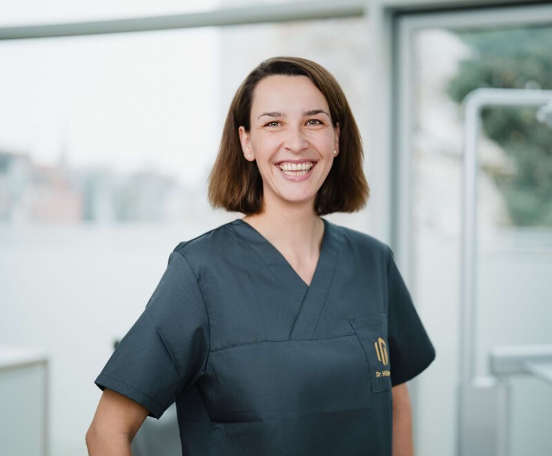 Freundliches Portrait von Dr. Anna H&uuml;ttmann in schwarz/grauem Kasack vor der Fensterfront eines Behandlungszimmers