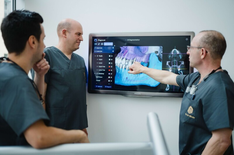 Die Experten der Praxis stehen vor einem gro&szlig;en Bildschirm und analyiseren den 3D-Scan eines Kiefers, um den EIngriff pr&auml;zise zu planen.
