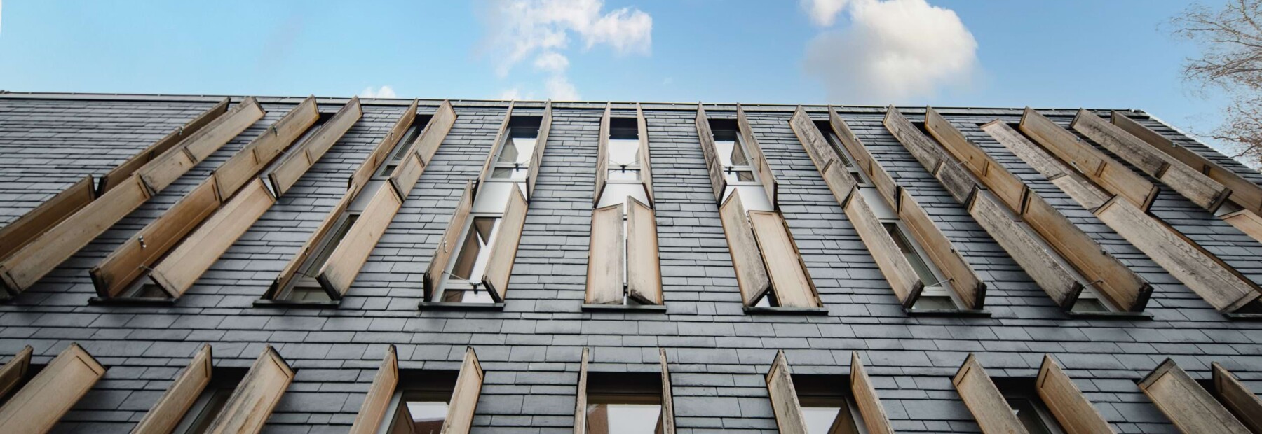 Au&szlig;enaufnahme des modernen Praixsklinik-Geb&auml;udes. Das Bild ist von unten nach oben aufgenommen und zeigt die mit Schiefern verkleidete Fassade und blauen Himmel im Hintergrund.
