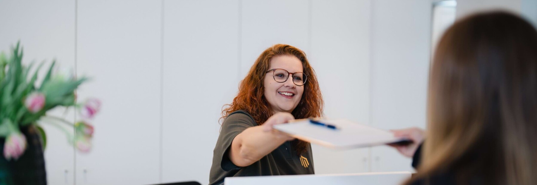 Eine freundliche Mitarbeiterin am Empfang der Praxis reicht eine Aufkl&auml;rungsmappe an eine Patientin, die hinter dem Tresen steht.