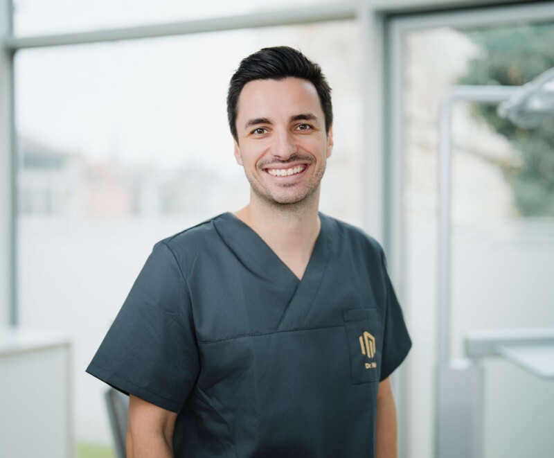 Freundliches Portrait von Dr. Alexander Mai in schwarz/grauem Kasack vor der Fensterfront eines Behandlungszimmers