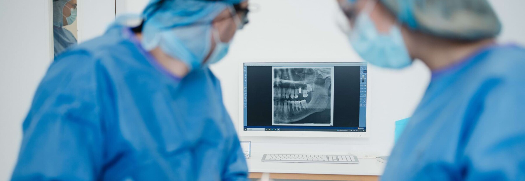 Dr. Will und Dr. Mai in OP-Kleidung w&auml;hrend einem Eingriff. Im Hintergrund ist ein Bildschirm mit der R&ouml;ntgenaufnahme eines Kiefers zu sehen.